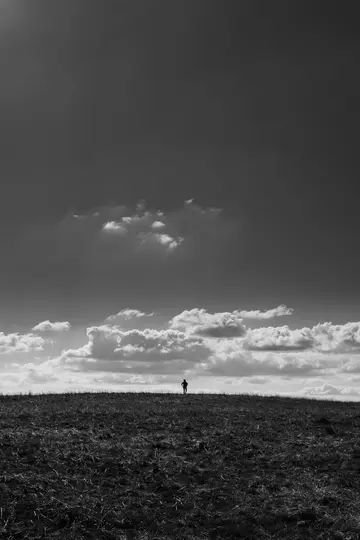 A black and white photo shows a tiny, silhouetted figure standing on the distant horizon of a grassy hill under a cloudy sky.