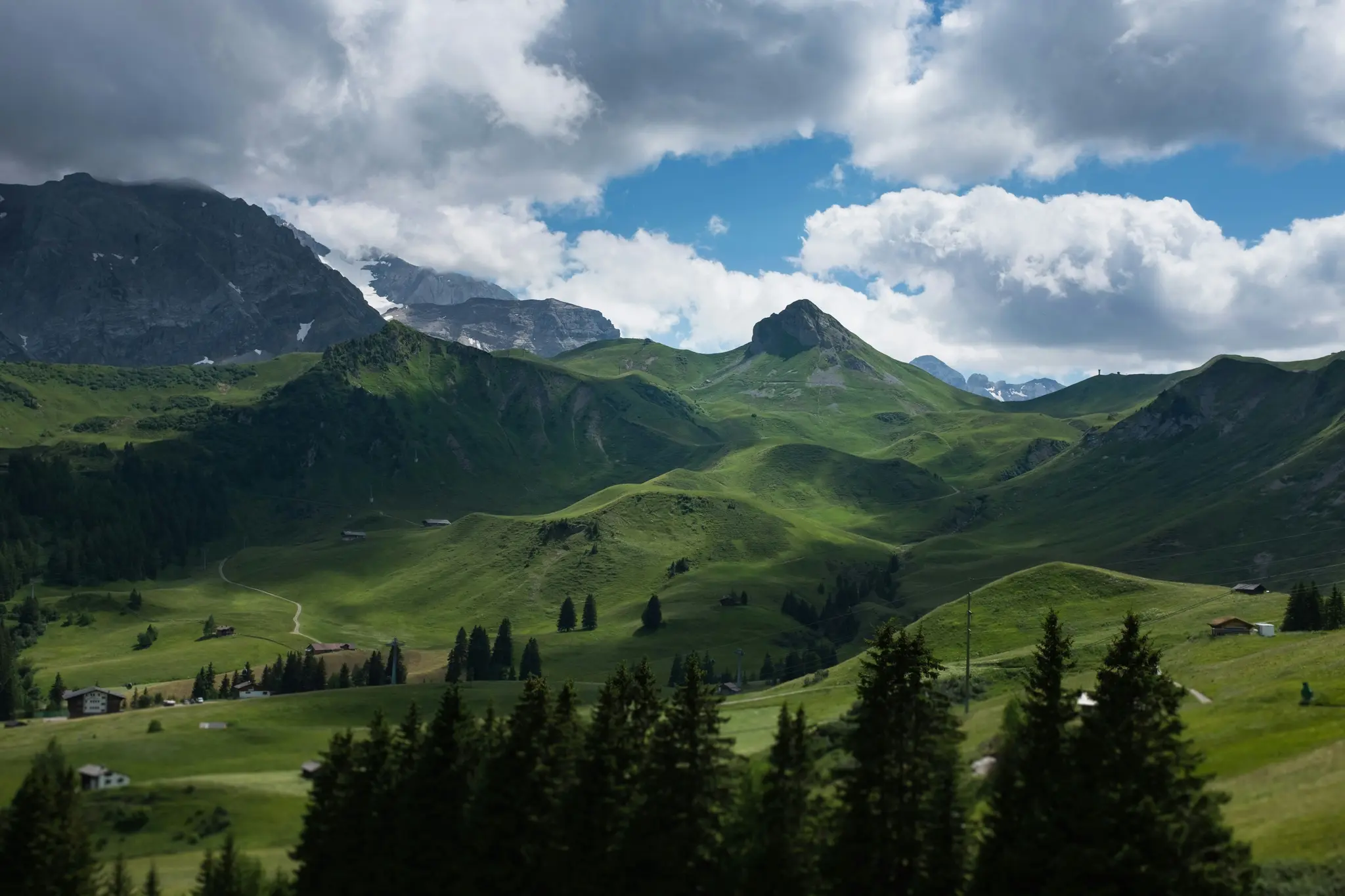 A panoramic view of lush green hills and valleys, with dark, snow-capped mountains in the background under a cloudy sky.