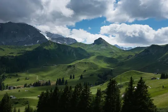 A panoramic view of lush green hills and valleys, with dark, snow-capped mountains in the background under a cloudy sky.