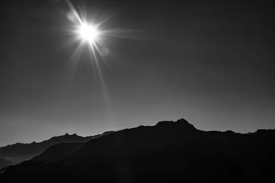 A black and white landscape features a brilliant sunburst in the upper left, casting rays over dark, silhouetted mountains.