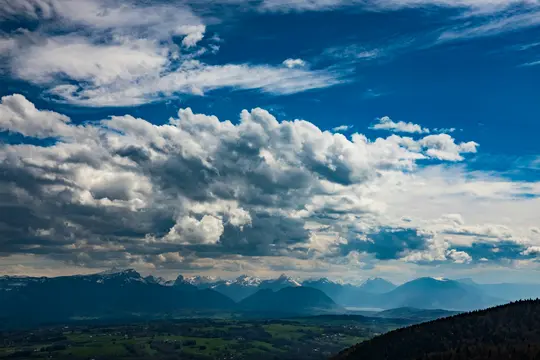 A wide landscape photo shows a valley with green fields and a dark forest in the foreground, leading to distant snow-capped…