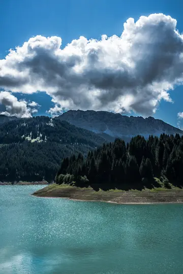 A vibrant blue-green lake reflects the sky, bordered by dark green pine forests and rugged mountains under a cloudy sky.