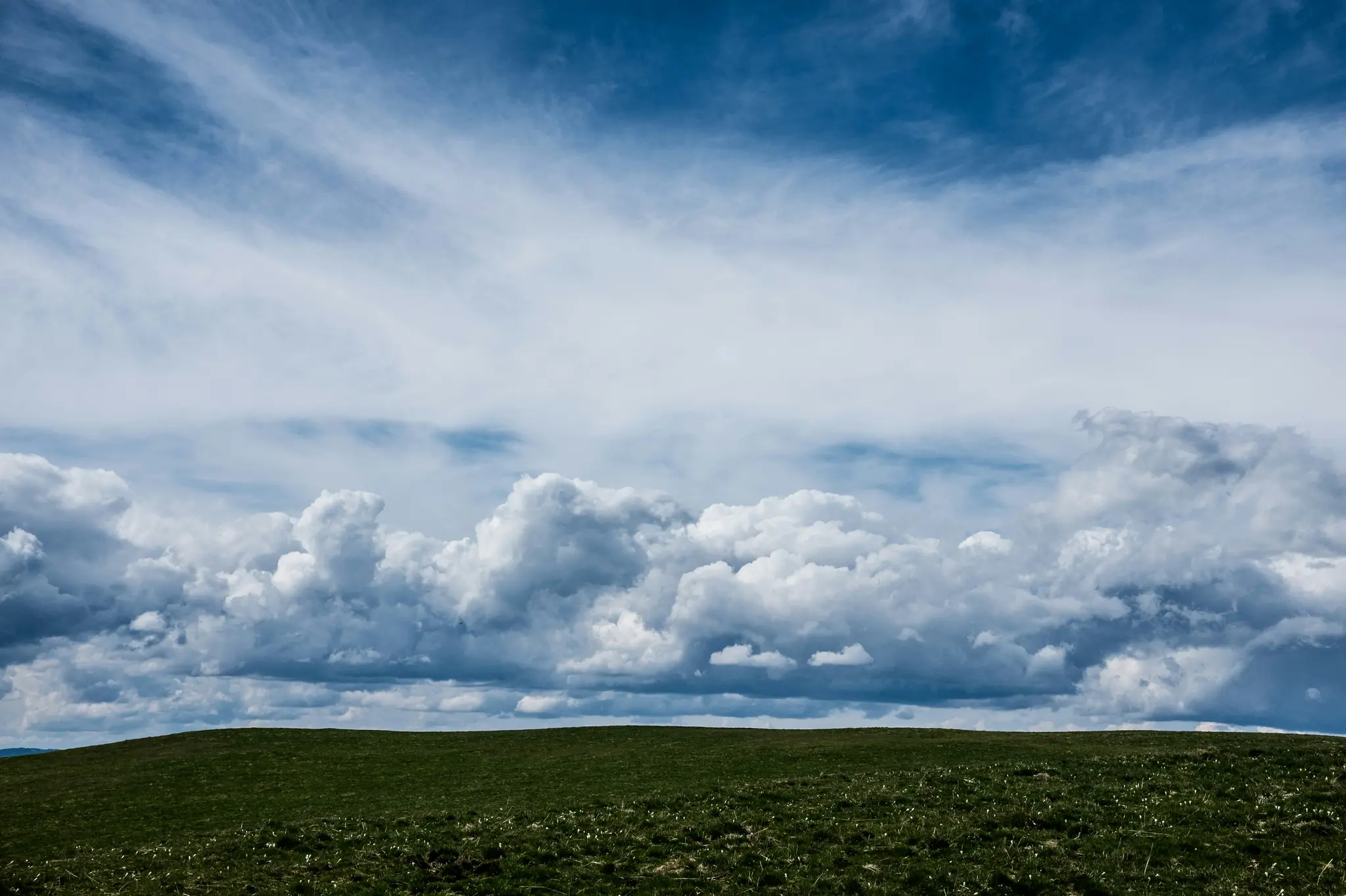 Et bredt landskabsfoto, der viser en mørkegrøn, græsfyldt bakke i forgrunden under en vid blå himmel med hvide og grå skyer.