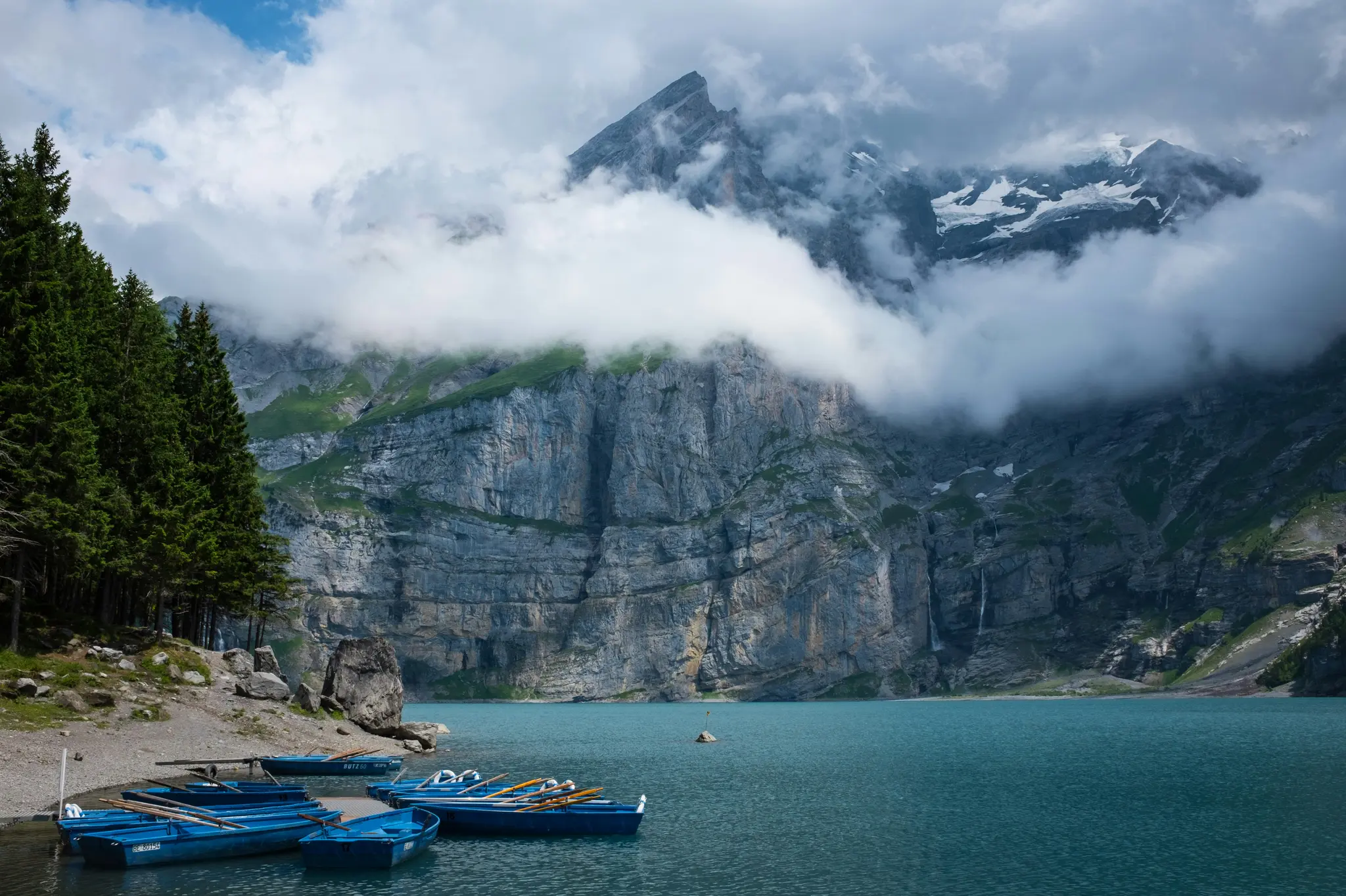 A wide landscape photo of a serene mountain lake with several blue rowboats docked on a rocky shore, framed by tall…