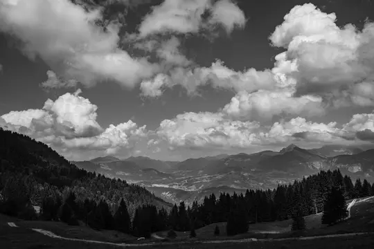 A dramatic black and white landscape features a vast valley nestled between forested mountains under a sky filled with…