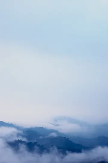 Vertikální krajinářská fotografie horských vrcholů zahalených v husté, měkké mlze a oblacích, vytvářející klidnou atmosféru.
