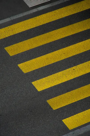 Et overset billede af et fodgængerfelt med kraftige gule striber malet diagonalt hen over mørkegrå asfalt. En lille…