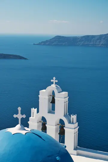 Ein weißer Glockenturm mit drei Glocken und einer blauen Kirchenkuppel im Vordergrund, vor einem tiefblauen Meer und in der