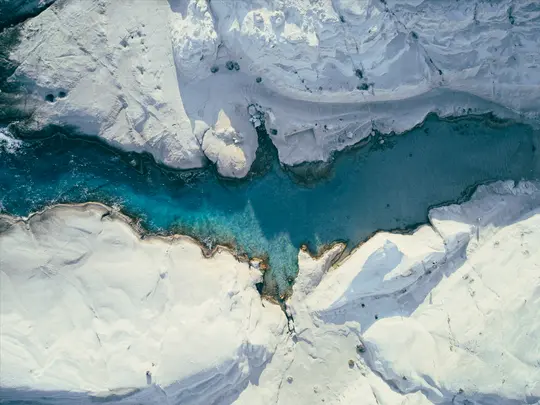 An aerial shot shows a vibrant blue river or inlet snaking through stark white rock formations, with clear water revealing…