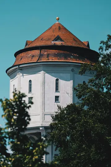 A white octagonal tower with a weathered red-tiled conical roof and a golden finial stands against a clear blue sky. Dark…