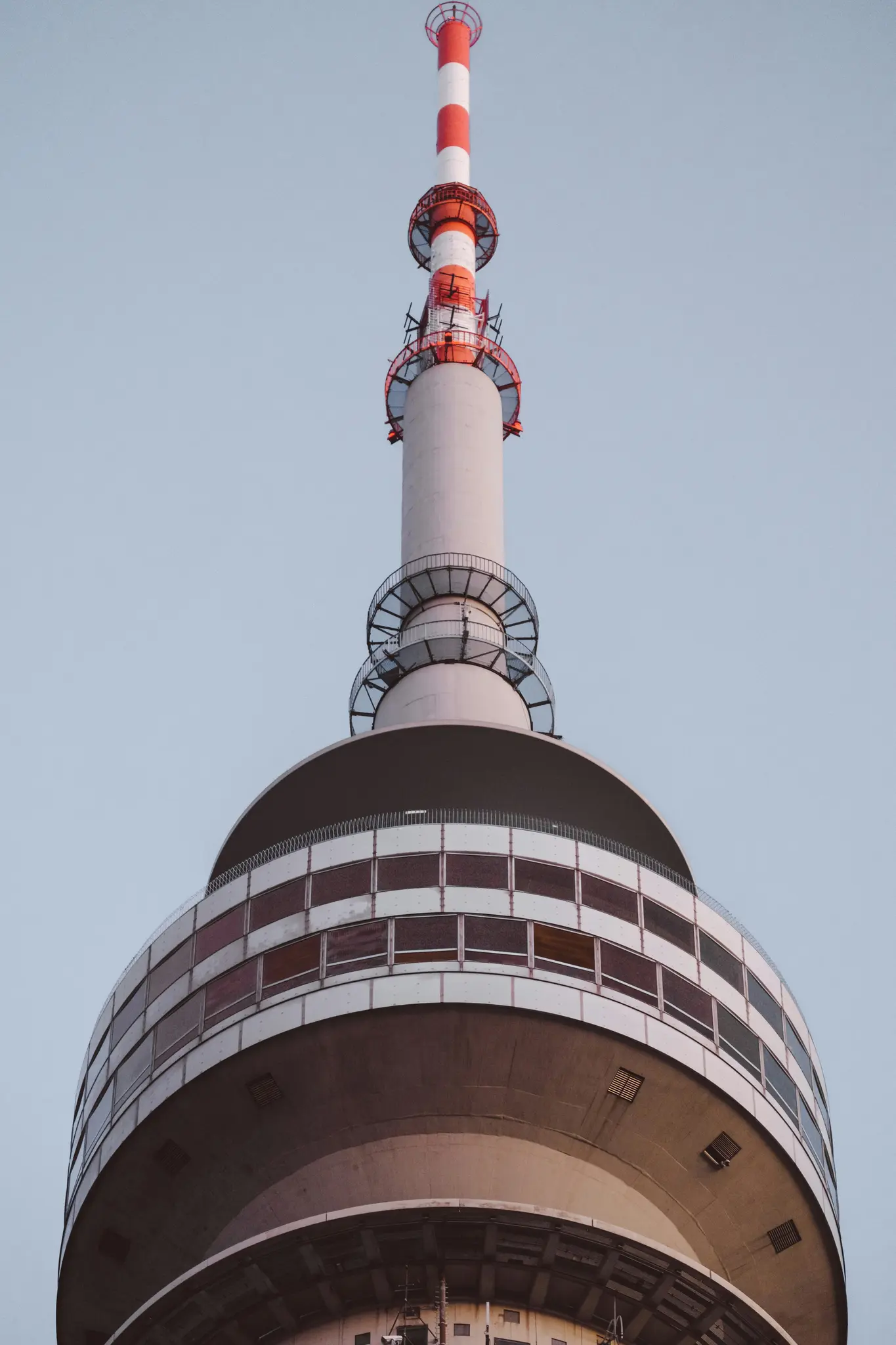 A close-up, low-angle view of a tall, modern telecommunications tower with a red and white striped antenna against a pale…