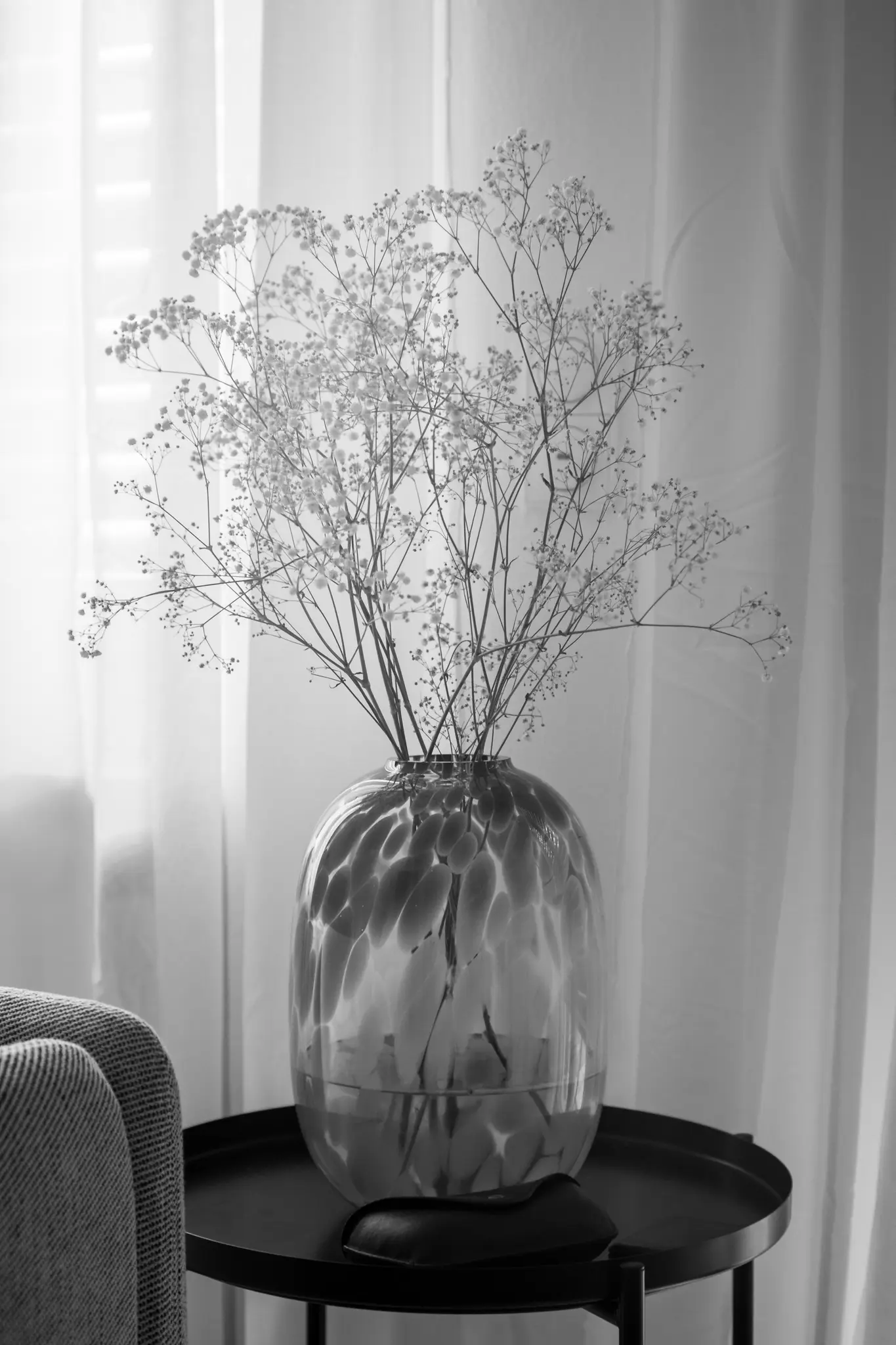 A black and white photo shows a glass vase with a spotted pattern holding dried baby's breath flowers on a dark round table.