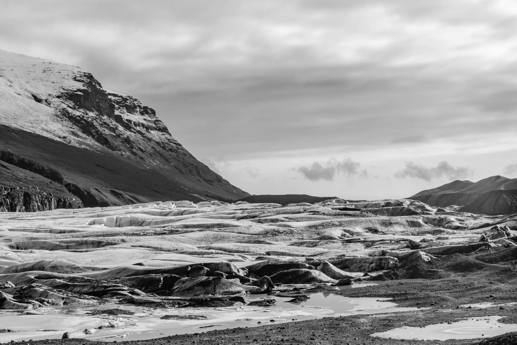 Et bredt sort-hvidt landskabsfoto af en gletsjer med et snedækket bjerg til venstre og en skyet himmel.