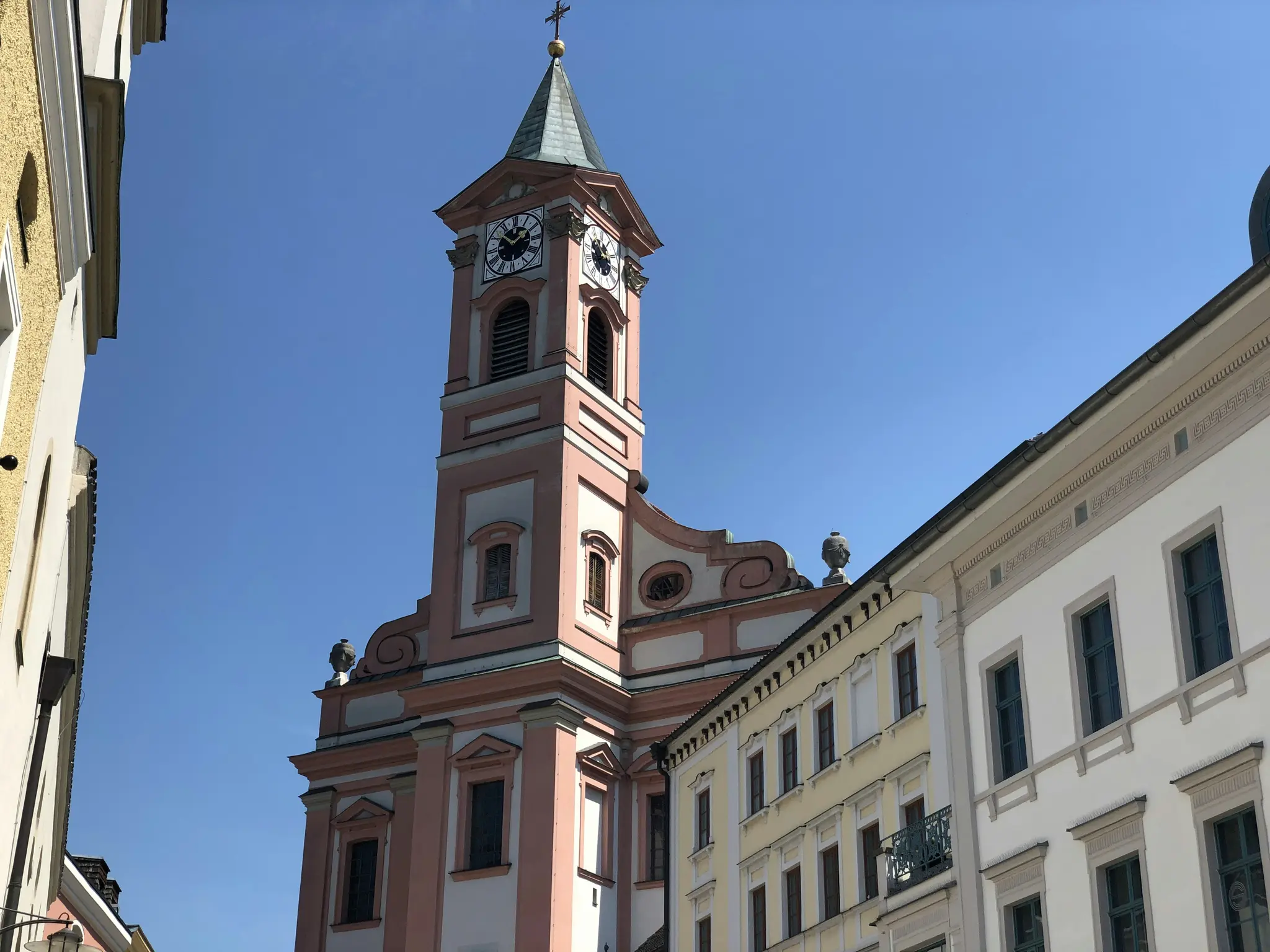 A low-angle view of a tall, pink church tower with a green spire and clock faces, set against a clear blue sky. Buildings…