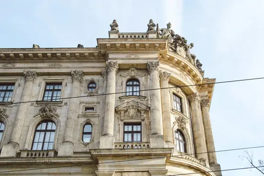 A low-angle shot of a grand, light-colored stone building featuring classical columns, arched windows, and intricate…