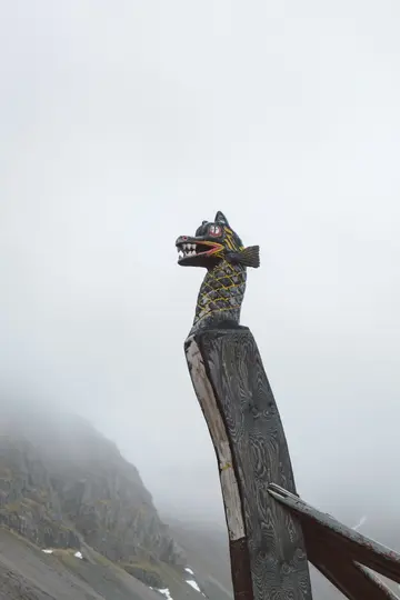 En detaljeret træskulptur af et dragehoved, malet sort, gult og rødt, på stævnen af et skib, mod en baggrund af…