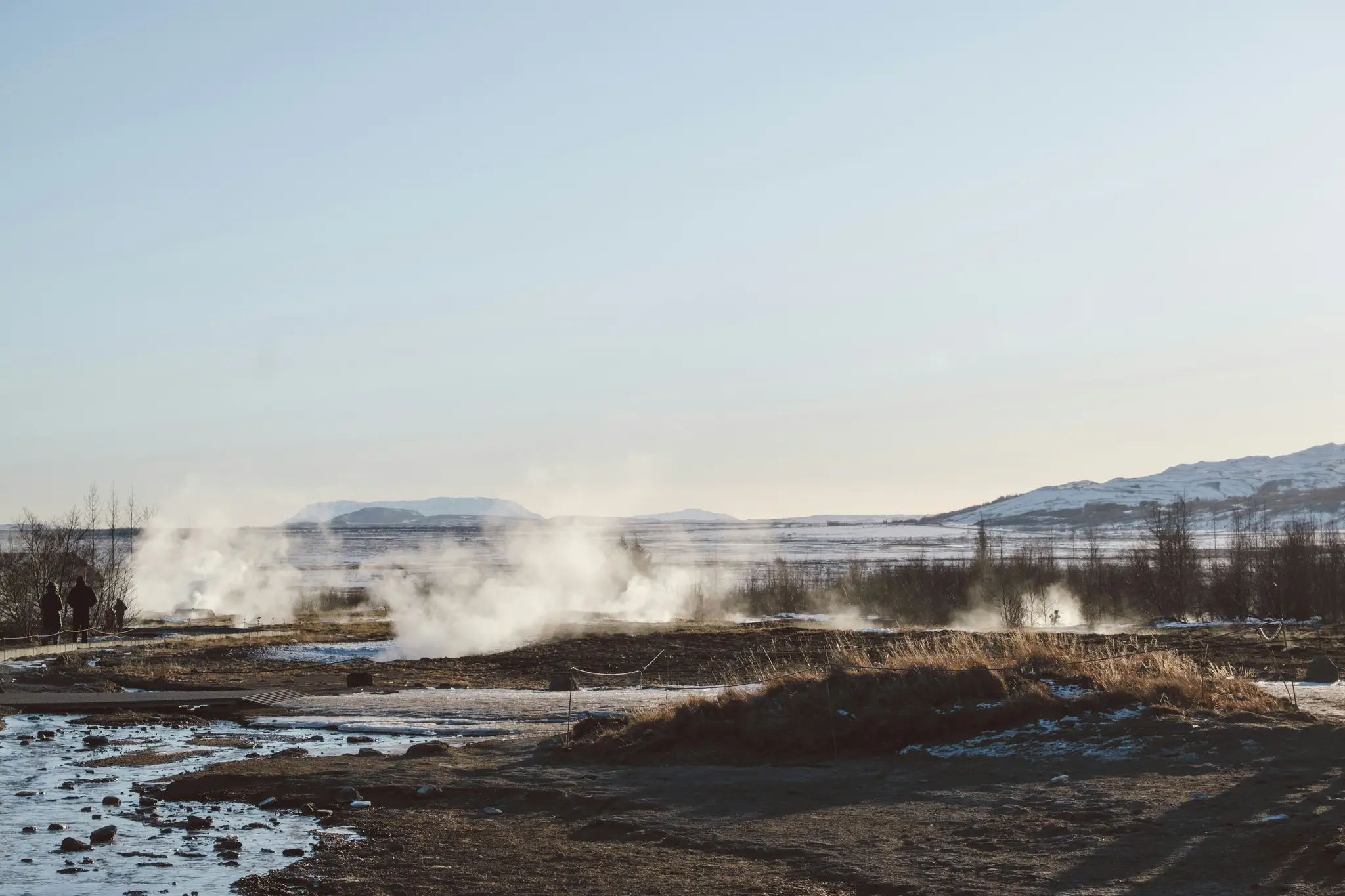 A wide landscape photo of a geothermal field in winter, with white steam rising from multiple spots on the brown ground.…