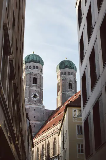 Et kig på de to ikoniske grønne spir på Frauenkirche i München, indrammet af de høje, moderne bygninger i en bygade.
