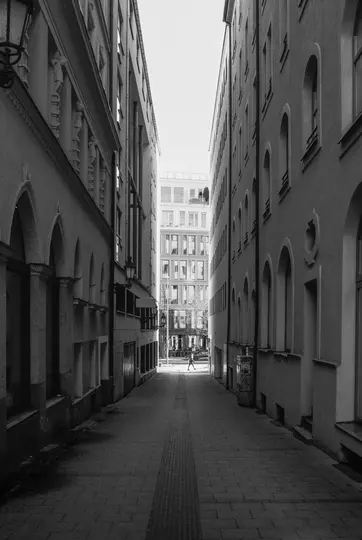 A black and white photo of a narrow alleyway flanked by tall buildings, with a lone figure walking towards a bright opening.