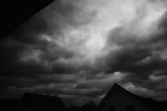 A black and white photo of a sky filled with dark, heavy clouds, with the silhouettes of house rooftops below.