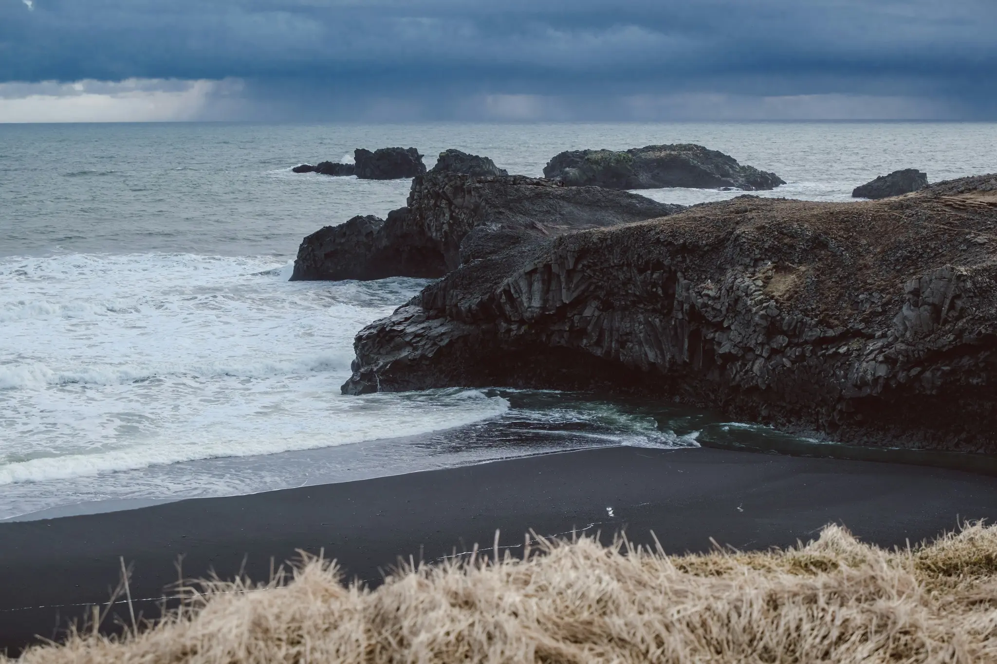 Et bredt skud af en mørk, stormfuld kystlinje. Bølger slår mod sorte vulkanske klippeformationer og en sort sandstrand.