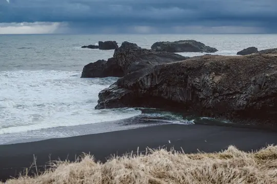 Et bredt skud af en mørk, stormfuld kystlinje. Bølger slår mod sorte vulkanske klippeformationer og en sort sandstrand.
