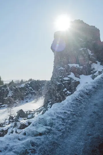A bright sun shines intensely from behind a large, snow-dusted rock formation on the right, illuminating a deep…