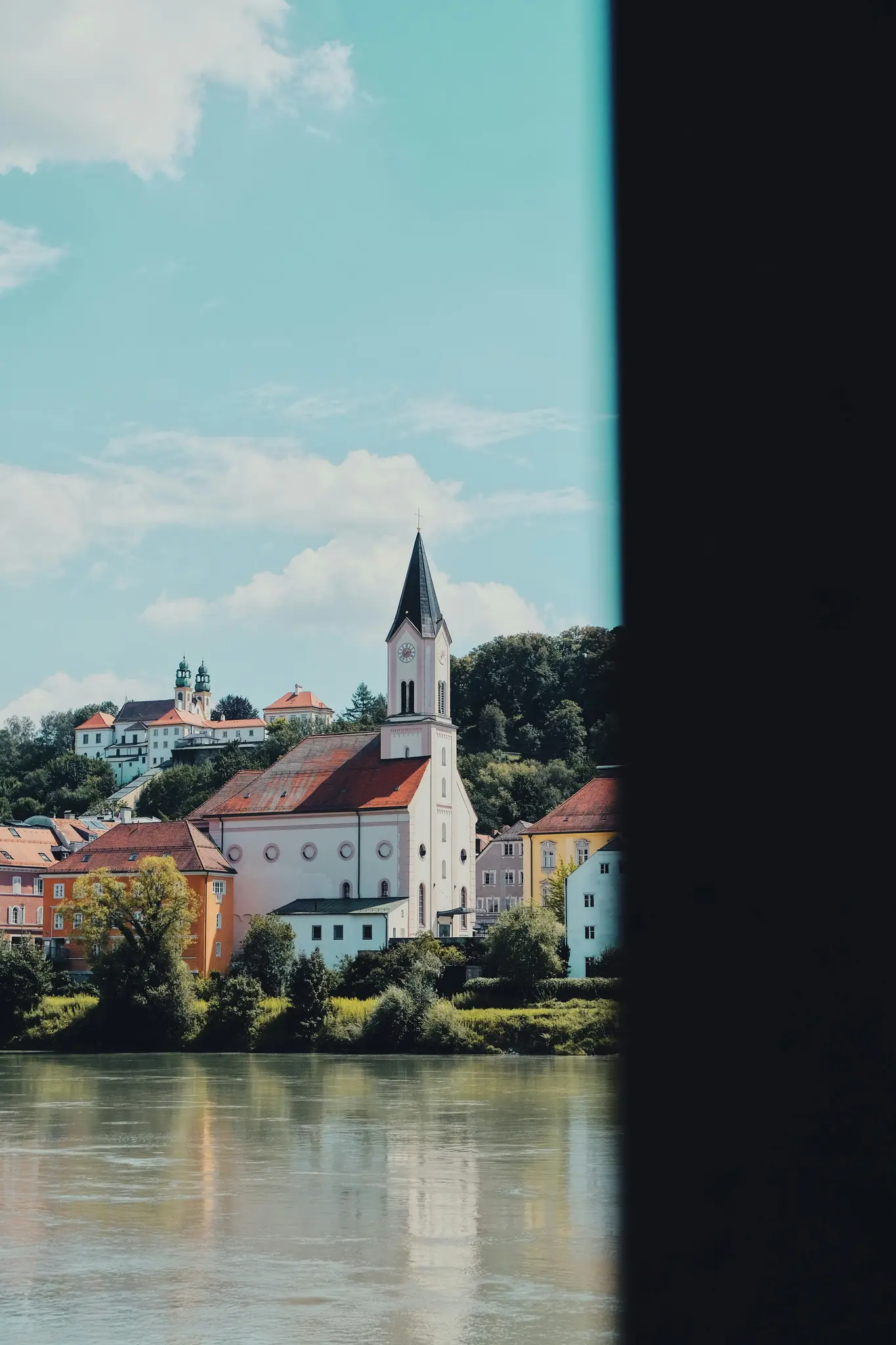 A white church with a black spire and red roof dominates a hillside town by a river, under a blue sky with clouds.