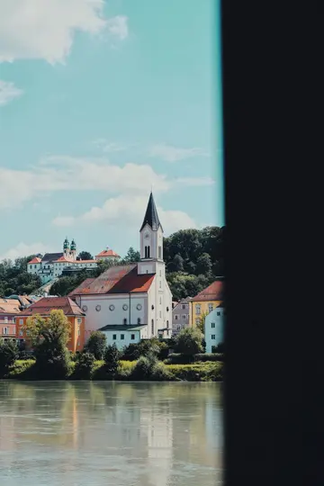 A white church with a black spire and red roof dominates a hillside town by a river, under a blue sky with clouds.