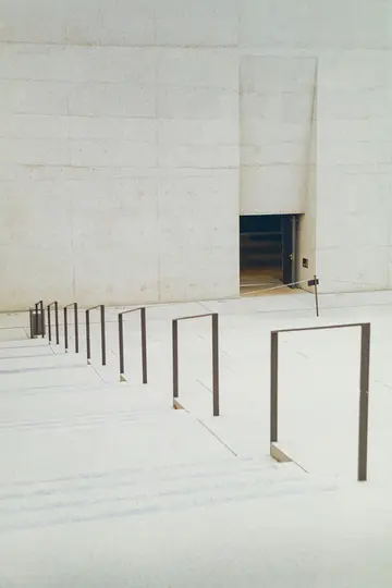 A minimalist photo of a light grey concrete building with a dark recessed entrance, next to a snow-covered ground with a…