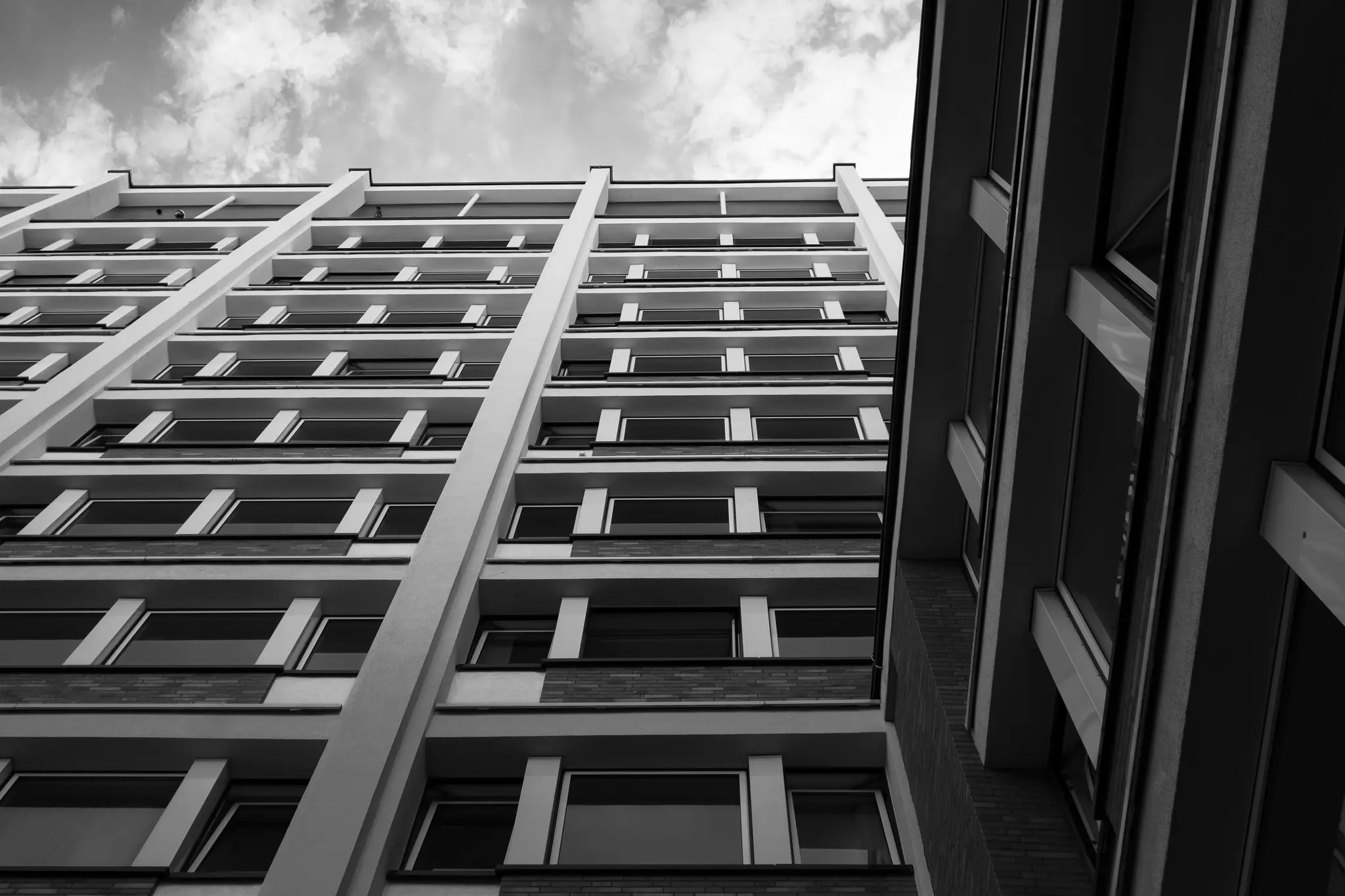 A black and white, low-angle shot of two tall buildings with many windows, viewed from below against a cloudy sky.