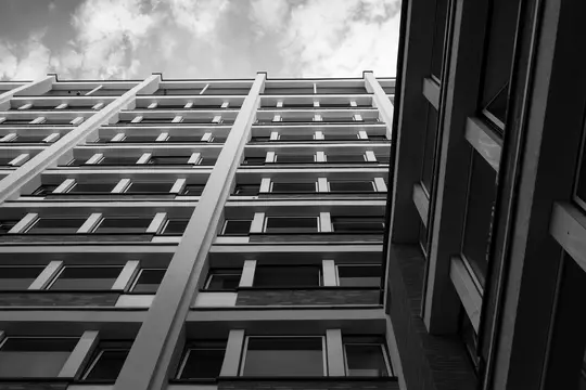 A black and white, low-angle shot of two tall buildings with many windows, viewed from below against a cloudy sky.