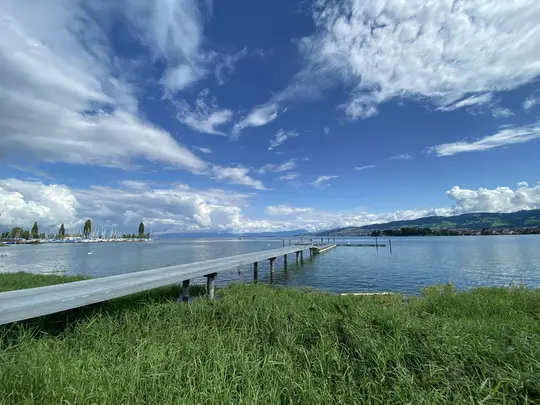 A long, grey pier stretches into a blue lake under a bright sky with fluffy white clouds. Green grass fills the foreground.