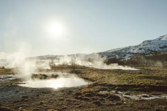 A wide landscape view of a geothermal field with steam rising from multiple hot springs and pools. The foreground features…