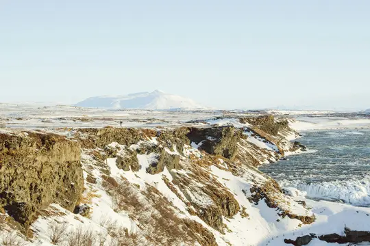 A wide shot of a snow-covered winter landscape. A lone figure stands on a rocky cliff overlooking a partially frozen river.