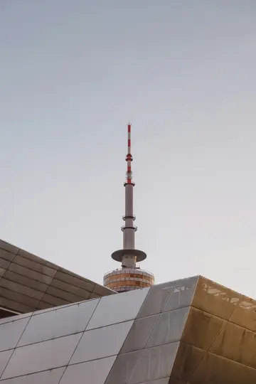 A tall, slender communication tower with a red and white striped antenna section, seen from a low angle above modern…