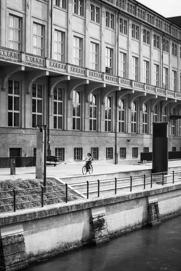 Et sort-hvidt billede viser en person på en cykel, der kører ad en sti ved siden af en kanal. En stor bygning med mange…