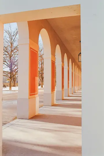 A long colonnade with cream-colored pillars and orange panels, featuring a series of arches that recede into the distance.…