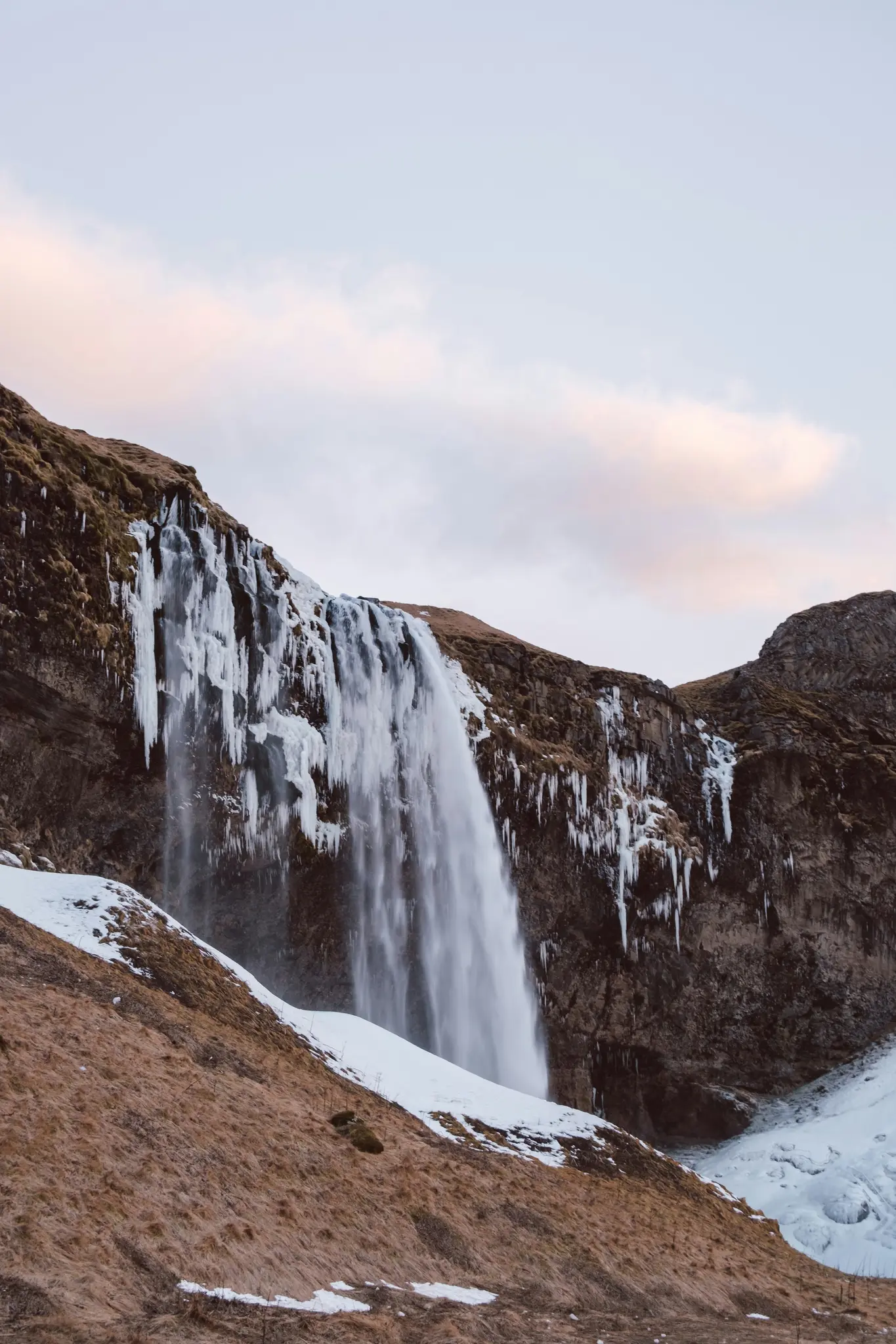 A tall waterfall plunges down a dark, mossy cliff face, partially covered in snow and long icicles. The foreground shows…