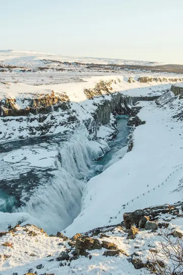 Et majestætisk vinterlandskab viser et kraftfuldt vandfald, der kaskaderer ned i en flod i en snedækket kløft. Gyldent lys…