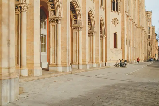 Ένα μακρύ, ανοιχτόχρωμο κτίριο με πολλά τοξωτά παράθυρα και κολώνες εκτείνεται κατά μήκος ενός πλακόστρωτου δρόμου. Ένα άτομο