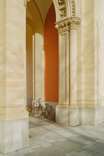 En sort cykel med brunt sæde er parkeret under en stenbuegang. Baggrunden viser buer og en orange væg.
