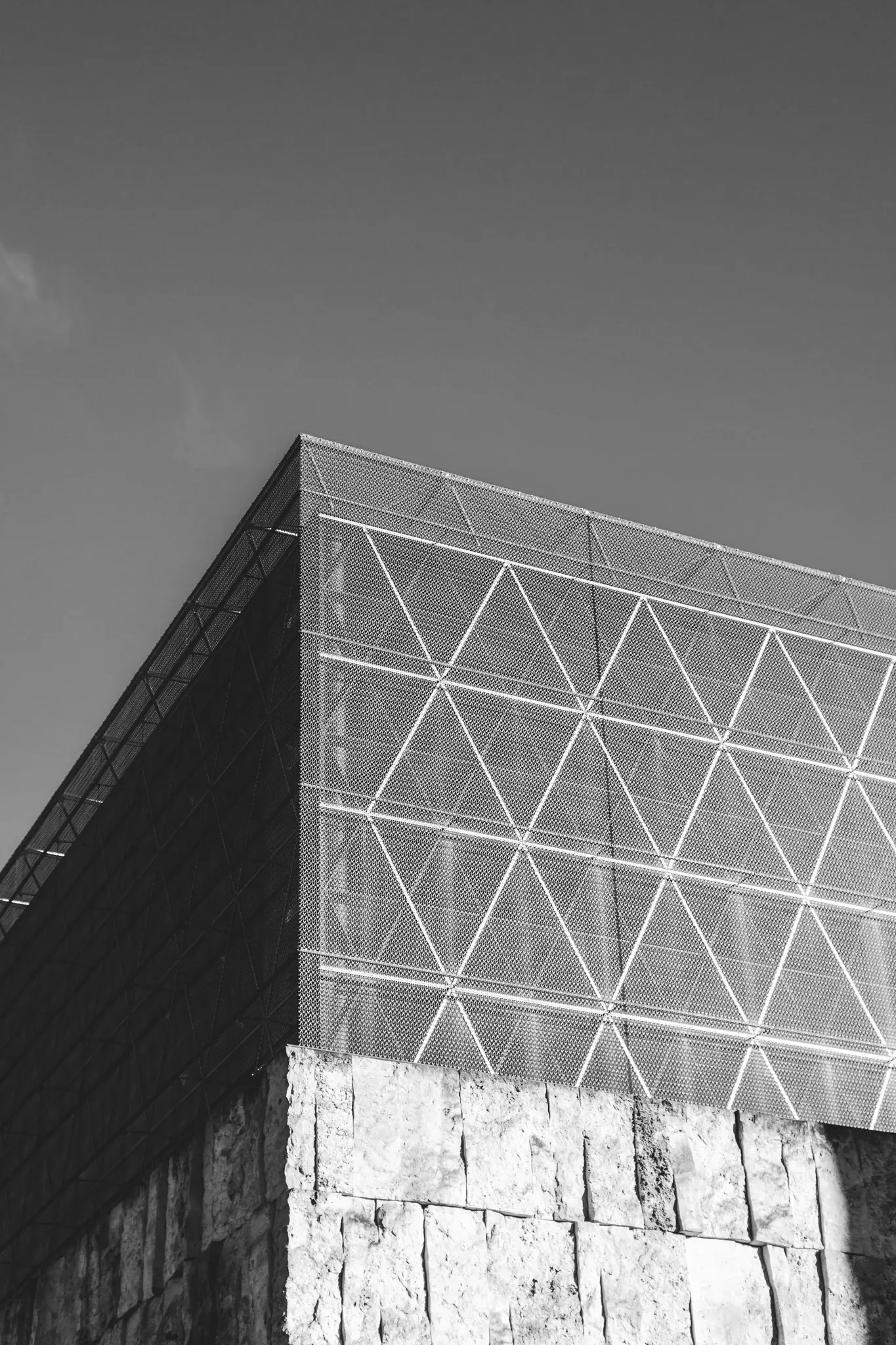A black and white photo of a modern building with a triangular patterned mesh facade above a rough stone wall.
