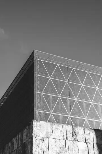 A black and white photo of a modern building with a triangular patterned mesh facade above a rough stone wall.