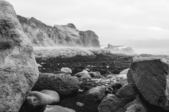 A black and white photo of a rocky beach. Large, textured rocks are in the foreground, with smaller pebbles and dark sand.