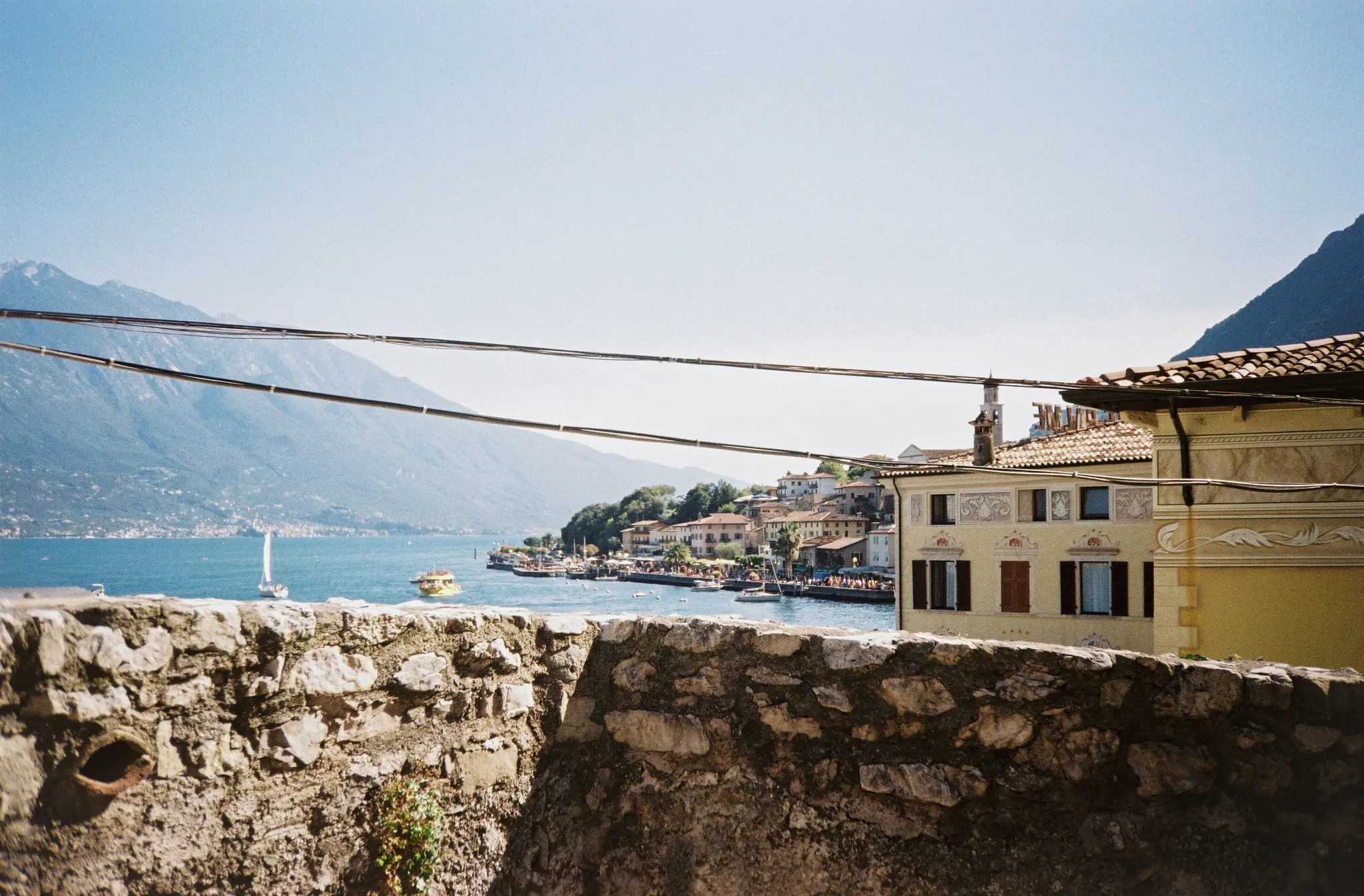 A wide shot of a lakeside town with mountains in the background, seen over a stone wall. Boats are on the water.