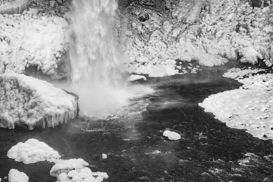 A black and white photo of a powerful waterfall plunging into a dark river, with thick snow and icicles on the surrounding…