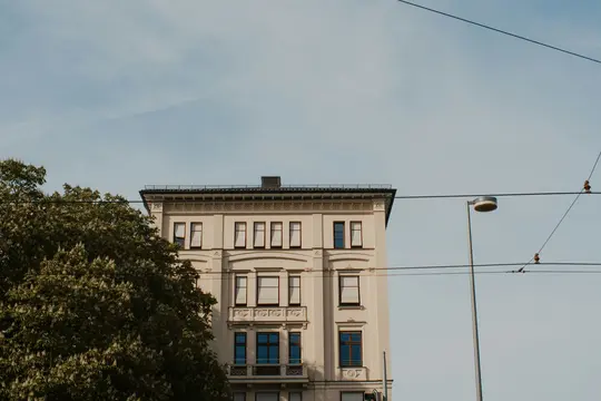 A cream-colored building with dark windows and ornate details stands under a pale blue sky, partially obscured by a green…