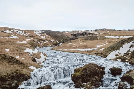 Et bredt landskabsfoto viser en flod med flere små vandfald, der løber gennem en dal. Brune bjergskråninger er dækket af…
