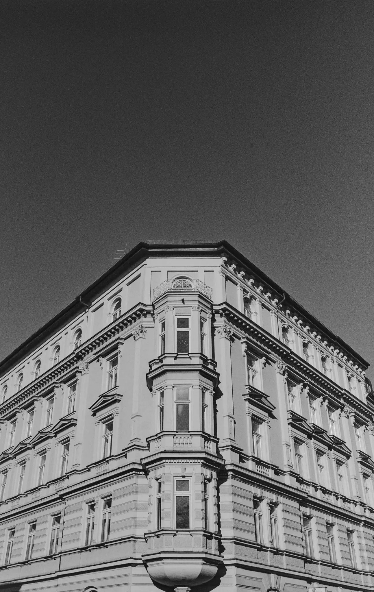 Nízký úhel, černobílá fotografie rohu staré budovy s arkýři, balkony a ozdobnými lištami.
